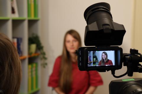 Na fotografii je žena v červené halence, která je natáčena při rozhovoru na kameru.